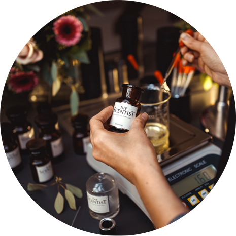 Zwei Hände mischen in einem Duftlabor Parfum. In der einen Hand wird eine braune Flasche mit dem Etikett „THE SCENTIST“ gehalten, in der anderen eine Pipette über einem Becherglas auf einer digitalen Waage. Im Hintergrund stehen weitere Fläschchen und Blumen.