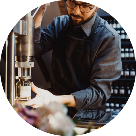 Ein Mann mit Brille und dunklem Hemd bedient eine Abfüllmaschine und verschließt ein Parfumfläschchen mit dem Label „THE SCENTIST“. Im Hintergrund stehen Regale mit weiteren Flaschen.