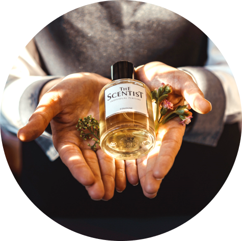 Zwei geöffnete Hände halten eine Parfumflasche mit der Aufschrift „THE SCENTIST – Individual Perfume“. In den Handflächen liegen zusätzlich kleine Blüten und grüne Kräuter. Die Szene ist warm beleuchtet.