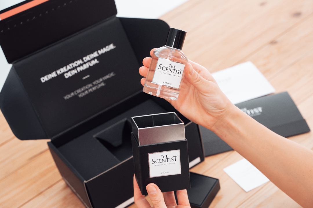 Zwei Hände halten ein Parfumfläschchen und die dazugehörige schwarze Box mit dem Etikett „THE SCENTIST – Individual Perfume“. Im Hintergrund liegt eine geöffnete Verpackung mit dem Aufdruck „DEINE KREATION. DEINE MAGIE. DEIN PARFUM.“ auf hellem Holztisch.